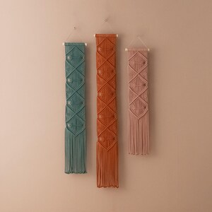 May include: Three macrame wall hangings with geometric patterns. The hangings are in shades of teal, orange, and pink.