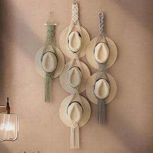 Puede incluir: Tres colgadores de pared de macramé que exhiben seis sombreros de paja de colores claros. El colgador izquierdo es verde salvia, el central es blanco roto y el derecho es gris. Cada colgador presenta nudos intrincados y flecos largos.