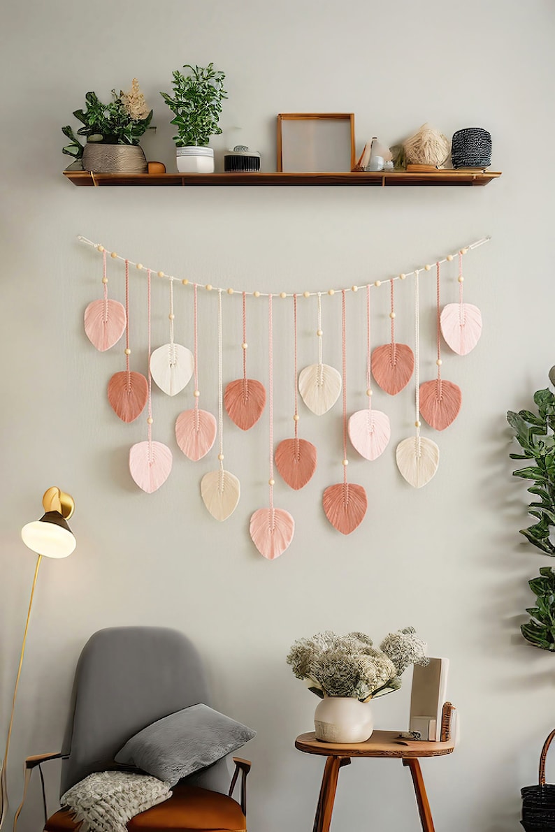 May include: A wall hanging made of woven yarn in the shape of leaves. The leaves are in shades of pink, cream, and brown. The leaves are strung together with wooden beads and twine. The wall hanging is hanging on a white wall.