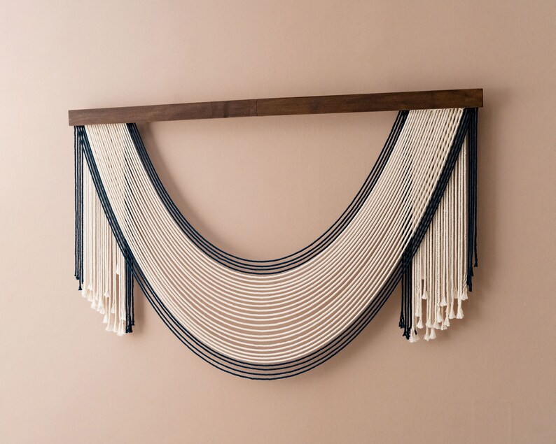 May include: A wall hanging made of white and dark blue yarn, with a wooden dowel at the top. The yarn is woven into a curved shape, creating a modern and minimalist design.