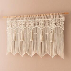 May include: Cream-colored macrame wall hanging. The piece features intricate knotted patterns, including geometric shapes and tassels. It is suspended from a natural wooden dowel, adding a touch of bohemian style to any space.