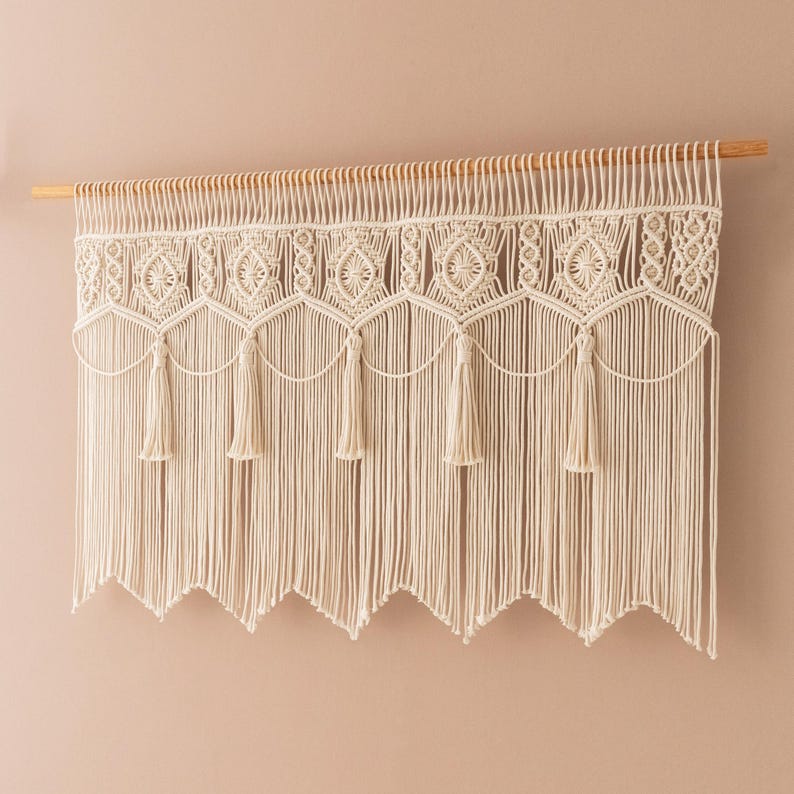 May include: Cream-colored macrame wall hanging. The piece features intricate knotted patterns, including geometric shapes and tassels. It is suspended from a natural wooden dowel, adding a touch of bohemian style to any space.