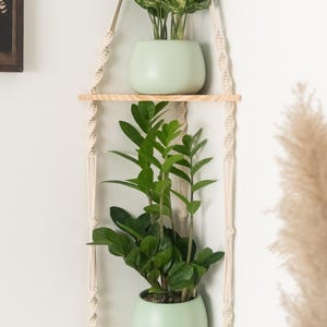 May include: A macrame plant hanger with two wooden shelves, each holding a potted plant. The top shelf has a plant with green and white leaves, while the bottom shelf has a plant with dark green leaves. The hanger is made of natural cotton rope and has a light wood finish.