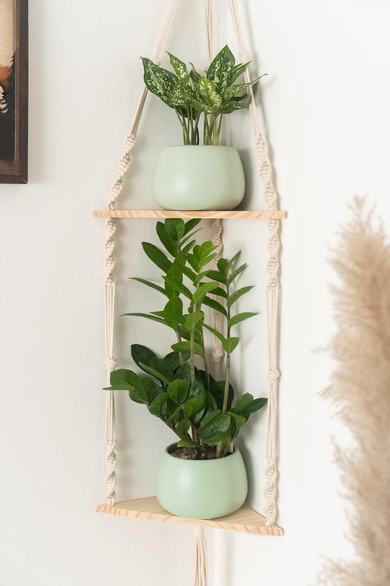 May include: A macrame plant hanger with two wooden shelves, each holding a potted plant. The top shelf has a plant with green and white leaves, while the bottom shelf has a plant with dark green leaves. The hanger is made of natural cotton rope and has a light wood finish.