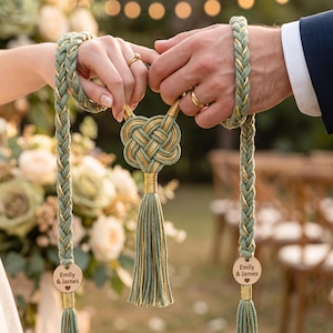 Könnte beinhalten: Ein Handfasting-Seil aus salbeigrünem und goldenem geflochtenem Seil mit einem zentralen gewebten Knoten und zwei runden Holzanhängern, die mit dem Text Emily & James und einem Herzsymbol graviert sind, abgeschlossen mit langen passenden Quasten.