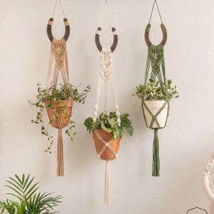 May include: Three macrame plant hangers with horseshoe accents. Each hanger holds a terracotta or white pot with greenery. The macrame is in beige, cream, and olive green, with a tassel at the bottom of each. The hangers are suspended against a white wall.