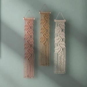 May include: Three macrame wall hangings with a leaf pattern. The hangings are in shades of beige, light pink, and white.
