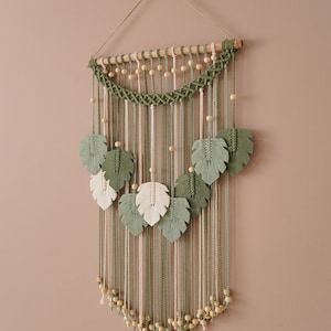 Op de afbeelding: Een groene en witte macram&eacute; wandhanger met houten kralen en bladvormen.