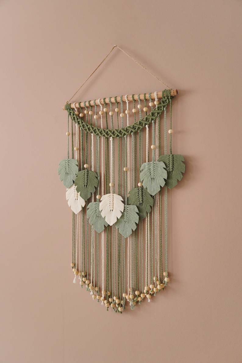 Op de afbeelding: Een groene en witte macram&eacute; wandhanger met houten kralen en bladvormen.