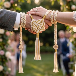 May include: A handfasting cord featuring braided cream and gold rope, a central woven knot, and two dangling tassels. Each tassel is accented with a circular wooden tag engraved with the text Emily & James. The cord wraps around two wrists.