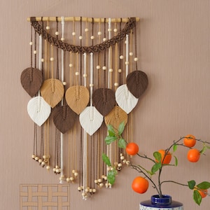 Op de afbeelding: Een bruine, beige en witte macram&eacute; wandhanger met houten kralen. De hanger heeft een geweven ontwerp met bladvormen. De hanger wordt getoond op een beige muur met een blauw-witte gepatroneerde vaas met oranje fruit.