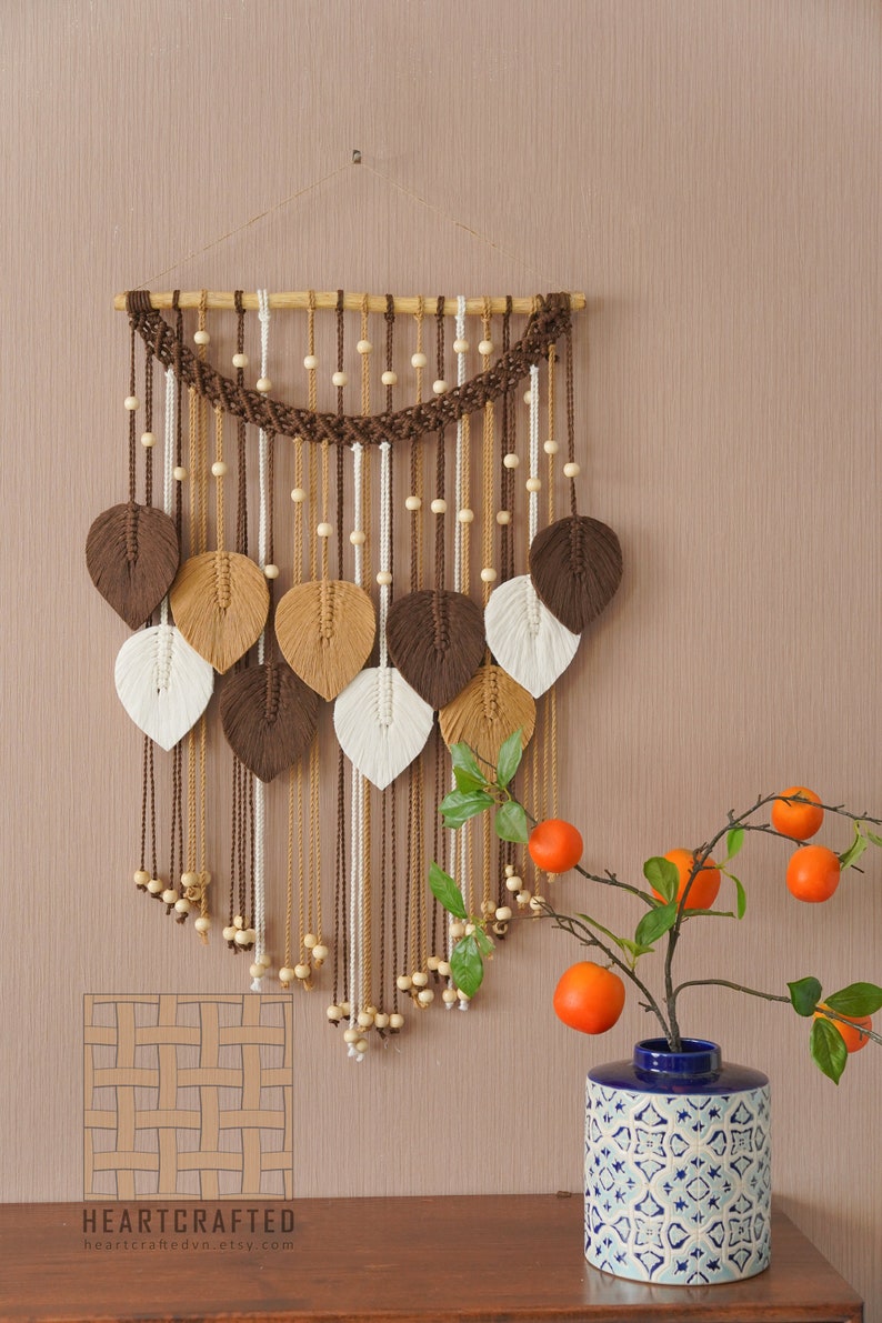 Op de afbeelding: Een bruine, beige en witte macram&eacute; wandhanger met houten kralen. De hanger heeft een geweven ontwerp met bladvormen. De hanger wordt getoond op een beige muur met een blauw-witte gepatroneerde vaas met oranje fruit.