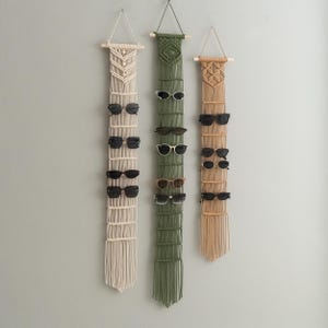 Könnte beinhalten: Drei Sonnenbrillen-Organizer aus Makramee in Creme, Olivgrün und Beige, jeweils mit einem Holzstab zum Aufhängen. Jeder Organizer bietet Platz für mehrere Sonnenbrillen. Die Sonnenbrillen sind schwarz und haben verschiedene Rahmenstile.