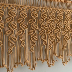 May include: A macrame wall hanging with a woven pattern and long fringe. The hanging is made of natural beige colored cord.