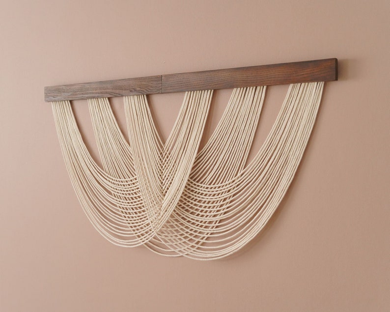 May include: A wall hanging featuring a horizontal dark brown wooden bar with multiple off-white cotton cords draped in overlapping, U-shaped loops. The cords are attached to the top wooden bar, creating a layered, cascading effect of curved lines.