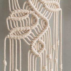 May include: A cream-colored macrame wall hanging featuring intricate knotted patterns. The design consists of thick cotton cord woven into symmetrical loops and geometric shapes, with long, vertical strands hanging down, each finished with a small knot.
