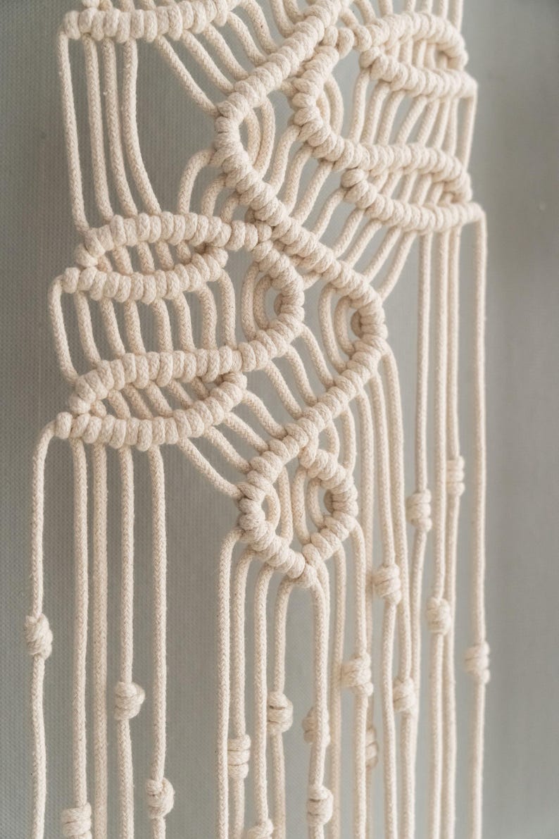 May include: A cream-colored macrame wall hanging featuring intricate knotted patterns. The design consists of thick cotton cord woven into symmetrical loops and geometric shapes, with long, vertical strands hanging down, each finished with a small knot.