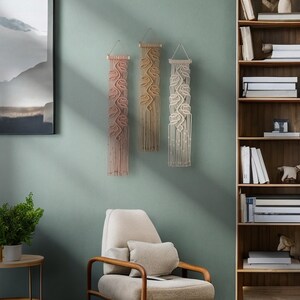 May include: Three macrame wall hangings with a leaf design in shades of pink, beige, and white. The hangings are made of cotton cord and are hung on a light green wall.