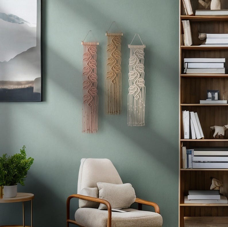 May include: Three macrame wall hangings with a leaf design in shades of pink, beige, and white. The hangings are made of cotton cord and are hung on a light green wall.
