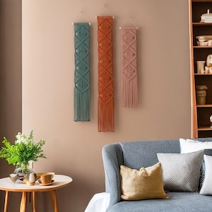 May include: Three macrame wall hangings in a living room setting. The hangings are made of natural fibers and have a geometric pattern. The hangings are in shades of teal, orange, and pink.