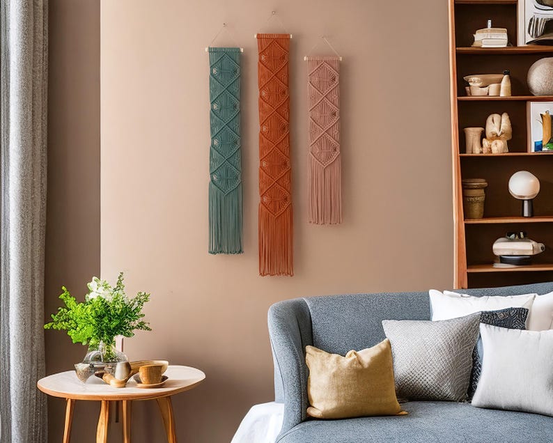 May include: Three macrame wall hangings in a living room setting. The hangings are made of natural fibers and have a geometric pattern. The hangings are in shades of teal, orange, and pink.