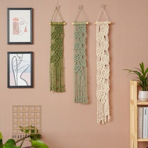 May include: Three macrame wall hangings with leaf designs. The hangings are made with natural cotton cord and are in shades of green and white. The hangings are hung on a light pink wall.