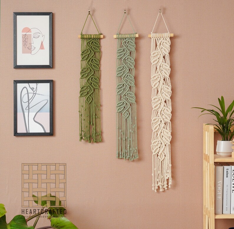 May include: Three macrame wall hangings with leaf designs. The hangings are made with natural cotton cord and are in shades of green and white. The hangings are hung on a light pink wall.