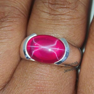 Może przedstawiać: Srebrny pierścionek z dużym, owalnym, różowym rubinem gwiaździstym. Pierścionek osadzony jest w prostej, srebrnej obrączce.