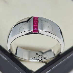 Może przedstawiać: Srebrna obrączka z rzędem czerwonych rubinów osadzonych w środku. Obrączka jest grawerowana napisem "Rounak Jewellers".