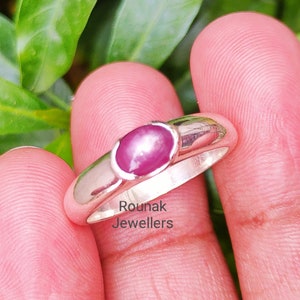 May include: A silver ring with a pink oval ruby gemstone. The ring is set in a simple band and has a polished finish. The text "Rounak Jewellers" is visible on the band.