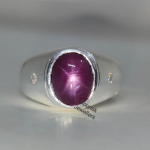 Peut inclure: Bague en argent avec une grosse pierre précieuse ovale rouge-violet. La pierre a un effet étoilé. De petites pierres claires sont serties de chaque côté de la pierre principale. Design classique.