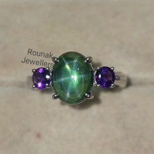 Peut inclure: Une bague en argent avec une pierre précieuse verte en forme d'étoile et deux petites pierres précieuses violettes. La bague est dans un écrin en velours rouge avec le texte "Rounak Jewellers" à l'intérieur.
