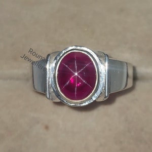 Peut inclure: Une bague en argent avec une pierre précieuse rouge ovale sertie dans une bélière en or. La bague a une bande blanche avec un motif décoratif.