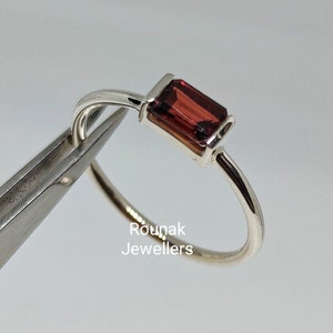 Peut inclure: Une bague en argent ornée d'une pierre précieuse rectangulaire rouge foncé. La bague a un fin anneau poli, et la pierre est sertie dans une lunette simple. La bague est tenue par une pince. Le texte "Rounak Jewellers" est visible.