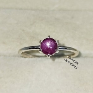 Peut inclure: Une bague en argent avec une pierre précieuse en rubis rouge. Le rubis présente un motif en forme d'étoile à l'intérieur. La bague est dans une petite boîte avec les mots "Rounak Jewellers" imprimés au fond.