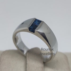 Op de afbeelding: Een zilveren bandring met drie kleine blauwe edelstenen in een kanaalsetting. De ring ligt op een witte displaystandaard. Rounak Jewellers is op de standaard gedrukt.