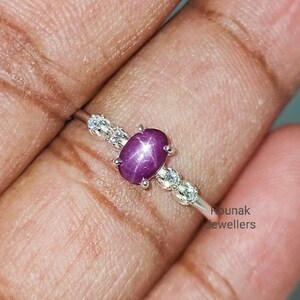 Peut inclure: Une bague en argent avec une pierre précieuse en rubis étoilé rose et de petites pierres précieuses claires sur la bande. La bague est portée sur un doigt.