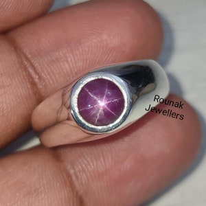 May include: A silver ring with a round, pink star ruby gemstone. The ring has a simple, rounded band. The text "Rounak Jewellers" is visible in the image.