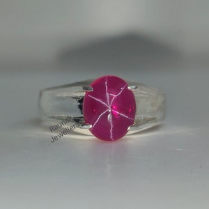May include: A silver ring with a red star ruby gemstone. The ruby is set in a bezel setting and has a star-shaped pattern.