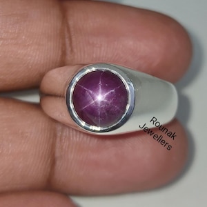 Può includere: Un anello d'argento con una pietra preziosa di rubino stella rosso. L'anello è incastonato in una fascia semplice con una parte superiore arrotondata. Il testo "Rounak Jewellers" è visibile sulla fascia.