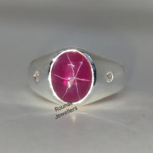 Könnte beinhalten: Ein silberner Ring mit einem ovalen, tiefroten Edelstein mit sternförmigem Muster. Der Ring hat eine glatte, polierte Oberfläche und kleine, klare Akzentsteine auf jeder Seite des Hauptsteins. Der Text "Rounak Jewellers" ist sichtbar.