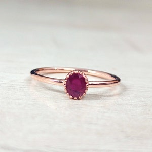 May include: A delicate rose gold ring with a single oval-shaped ruby gemstone set in a beaded bezel.