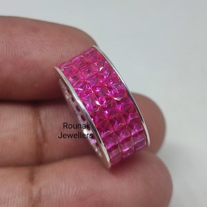 Puede incluir: Un anillo de plata con una banda ancha engastada con varias filas de piedras cuadradas de color rosa. El anillo está en una mano, y el texto "Rounak Jewellers" es visible en la banda.