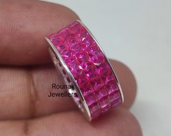 May include: A silver ring with a wide band set with multiple rows of pink square-shaped stones. The ring is on a hand, and the text "Rounak Jewellers" is visible on the band.