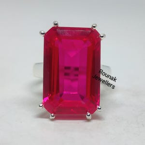 May include: A silver ring featuring a large, rectangular, bright pink gemstone. The gemstone is set with small silver prongs at each corner. The text "Rounak Jewellers" is visible on the right side of the image.