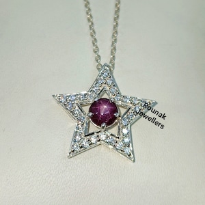 May include: A silver star-shaped pendant necklace with a red ruby gemstone in the center. The star is surrounded by smaller clear gemstones. The necklace is on a white background. Rounak Jewellers is written in the bottom right corner.