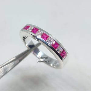 May include: A silver ring with alternating square-cut pink and clear gemstones. The ring is held by tweezers, with the text "Rounak Jewellers" visible on the side. The gemstones are set in a channel setting.