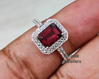 May include: A silver ring with a red gemstone surrounded by smaller white gemstones. The ring is on a finger.