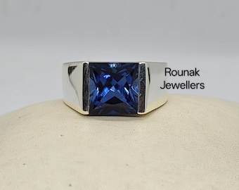 May include: A silver ring with a large, square, deep blue gemstone. The ring has a simple, modern design with a thick band. The text "Rounak Jewellers" is visible on the side of the ring.