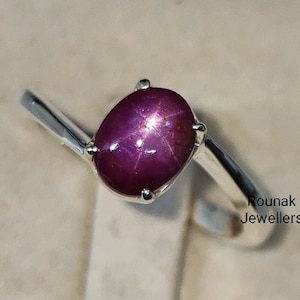Peut inclure: Une bague en argent avec une pierre précieuse en rubis étoilé ovale de couleur rouge foncé. Le rubis présente un motif en forme d'étoile au centre. La bague est posée sur un fond blanc. Rounak Jewellers.